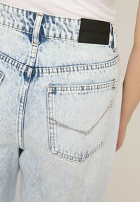 Jasnoniebieskie dżinsy z denimu o wyblakłym wykończeniu. Posiadają czarną skórzaną naszywkę w pasie oraz klasyczne tylne kieszenie z minimalnymi detalami przeszycia.