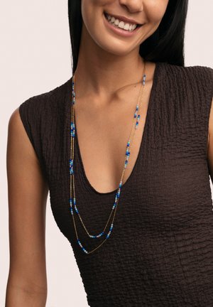 Gold chain necklace with multiple strands featuring small blue and white beads, worn against a textured dark brown top.
