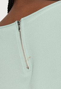 Haut vert clair avec une fermeture éclair métallique au dos. Le tissu a une finition texturée avec une petite tache visible près de la fermeture.