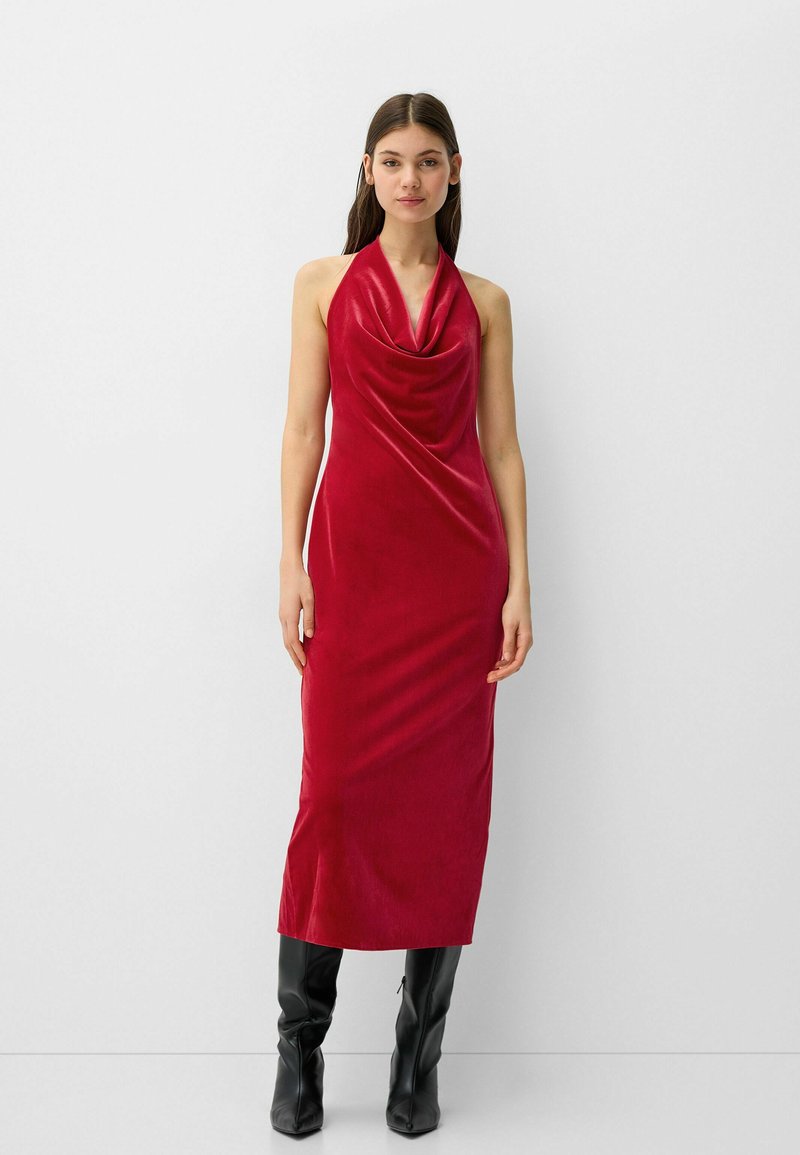 Robe rouge en velours à col halter, avec un décolleté en drapé, longueur au sol et texture lisse, assortie à des bottes noires jusqu'aux genoux.