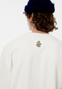 Bílá oversize mikina s texturovanou nášivkou s motivem naskládaných kamenů na zádech. Měkká bavlněná látka, volný střih.