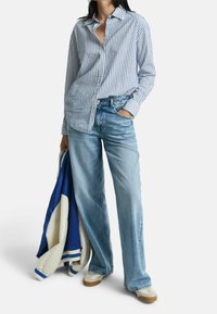 Blauw-wit gestreepte overhemd met knopen, losse pasvorm, gedragen met lichtblauwe wijde jeans. Houdt een blauw en crèmekleurig jack vast. Casual schoeisel.