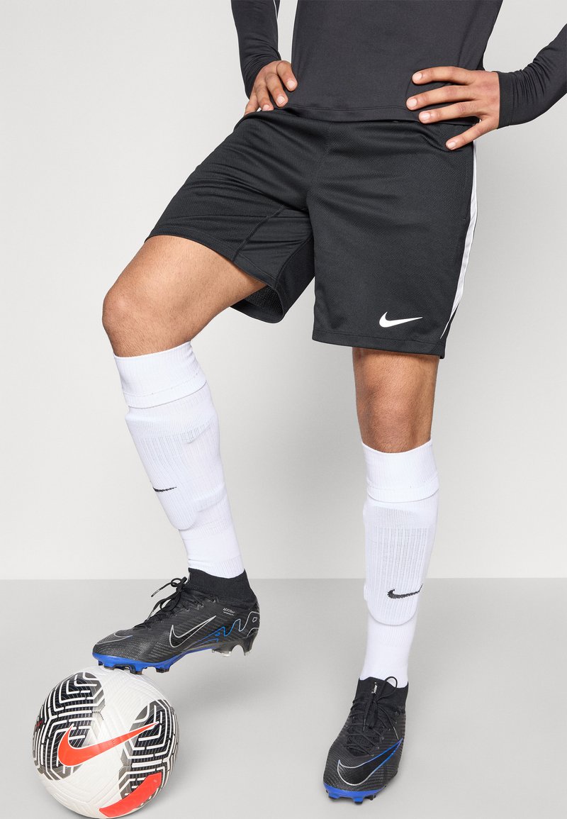 Schwarze Nike-Fußballshorts mit weißen Seitenstreifen, kombiniert mit weißen Kniestrümpfen. Schwarze Fußballschuhe mit blauen Akzenten und ein gemusterter Fußball.