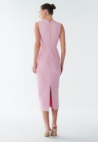 Frau in einem ärmellosen pinkfarbenen Midikleid mit Reißverschluss auf der Rückseite und Schlitz, trägt durchsichtige High-Heel-Sandalen vor weißem Hintergrund.