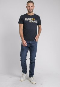 T-shirt bleu marine avec un texte graphique blanc et orange "BLUE 89 JEANS." Associé à un jean bleu foncé et des baskets blanches. Ajustement standard.