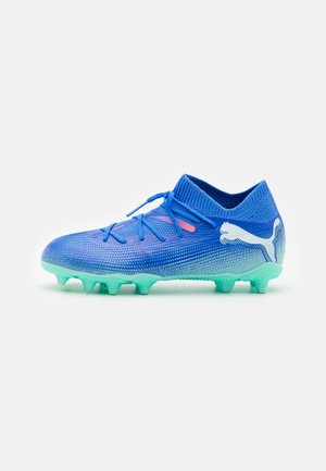 Botas de fútbol atléticas azules con parte superior de punto texturizado, acento rosa y tachuelas de goma verde para agarre. Logo de PUMA en el lado exterior.