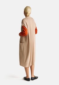 Béžový dlouhý vest, vyrobený z měkkého materiálu, se širokými průramky, bočními kapsami a žebrovaným lemem. Pevné černé boty doplňují celý outfit.