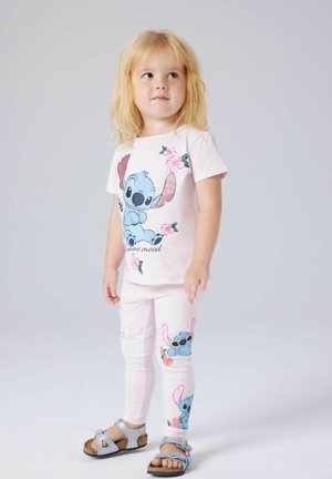 Blondes Kleinkind steht und schaut nach oben, trägt ein hellrosa Outfit mit einem blauen Cartoon-Charakter und Blumen, silberne Sandalen auf grauem Hintergrund.