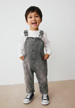 Niño pequeño sonriendo, vistiendo peto de mezclilla gris, camiseta blanca de manga larga y zapatillas negras, de pie contra un fondo claro y sencillo.
