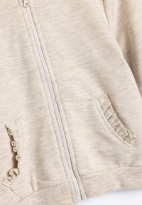 Beige Zip-Hoodie aus strukturiertem Stoff mit gerüschten Taschenrändern und Rippenbündchen am Saum und an den Handgelenken.