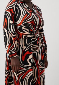 Vestido de manga larga con un lazo delantero, que presenta un atrevido patrón abstracto en negro, rojo y crema sobre una tela suave. Diseño de cuello tipo botón.