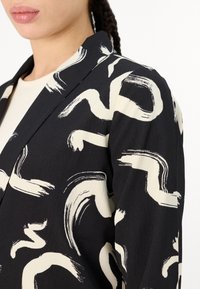 Mustalineblazer mustriga kreemja pintsliga mustriga. Struktureeritud disain, millel on lõige krae ja sile tekstuur.