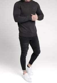 Mężczyzna w dopasowanym czarnym swetrze, przetartych czarnych skinny jeansach, w białych trampkach, z widocznymi tatuażami na rękach i kostkach, stojący na białym tle.
