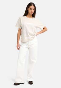 Μπεζ βαμβακερό μπλουζάκι με λευκό γραφικό σχέδιο και το κείμενο "ΑΝΟΙΚΤΟ ΜΥΑΛΟ." Συνδυασμένο με λευκά τζιν παντελόνια φαρδιάς γραμμής και μαύρα παπούτσια.