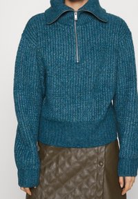 Pull en tricot bleu-vert avec un col montant et une fermeture éclair argentée, doté de poignets et d'un ourlet côtelés. Porté par-dessus une jupe en cuir marron matelassée.