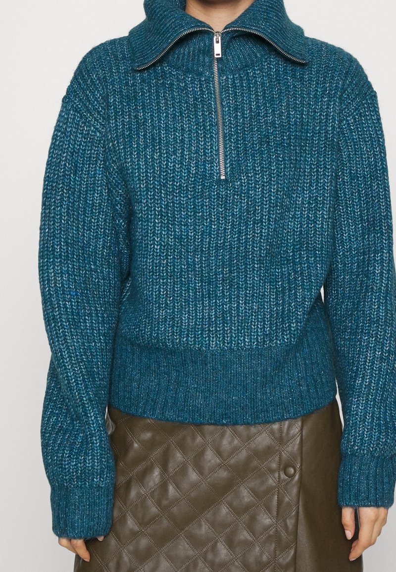 Pull en tricot bleu-vert avec un col montant et une fermeture éclair argentée, doté de poignets et d'un ourlet côtelés. Porté par-dessus une jupe en cuir marron matelassée.