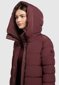 Burgundia värvi puffer-jakk suurte kapuutsiga, millel on lukk ja nööbid, ning tikitud disain. Varrukal on nööbi aksessuaar.