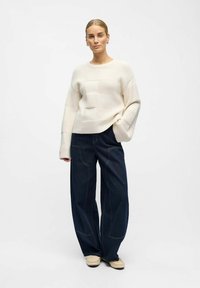 Suéter de punto crema con cuadros texturizados, combinado con pantalones de mezclilla oscuros de pierna ancha que presentan detalles de costura en contraste. Ajuste suave y relajado.
