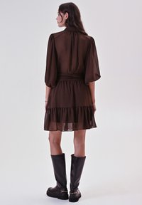 Donna che indossa un vestito trasparente marrone, lungo fino al ginocchio, con maniche a sbuffo e stivali neri fino al ginocchio, in piedi di spalle contro uno sfondo semplice.