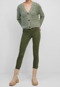 Grüner strukturierten Strickcardigan mit V-Ausschnitt und Knopfverschluss, kombiniert mit figurbetonten olivgrünen Cordhosen und beigen Ankle Boots.