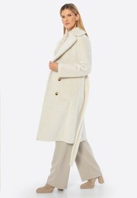 Abrigo crema de doble botonadura con cuello de muesca, textura suave y dos botones dorados. Llevado sobre pantalones beige y zapatos de tacón cuadrado marrones.