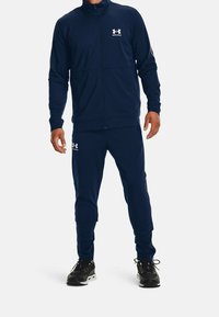 Mörkblå träningsdräkt gjord av slätt material. Jacka med full dragkedja och hög krage, byxor med elastisk midja. Har logotypaccenter i vitt.