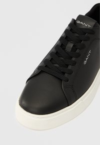 Črni usnjeni športni copat z belo gumijasto podplatom, ravnimi vezalkami in označbo blagovne znamke na strani. Ima gladko teksturo z zaobljenim prstnim delom.