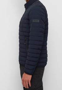 Marineblauw doorgestikt jack met een opstaande kraag, een zijzak met rits en een patch-logo op de mouw. Gladde textuur en lichtgewicht ontwerp.