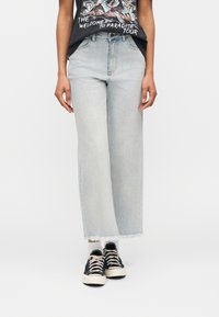 Άτομο που φοράει ανοιχτό μπλε τζιν παντελόνι με φαρδύ κόψιμο και κομμένο στο κάτω μέρος, μαύρα αθλητικά παπούτσια με λευκά κορδόνια, λευκές κάλτσες και μαύρο μπλουζάκι με σχέδιο.