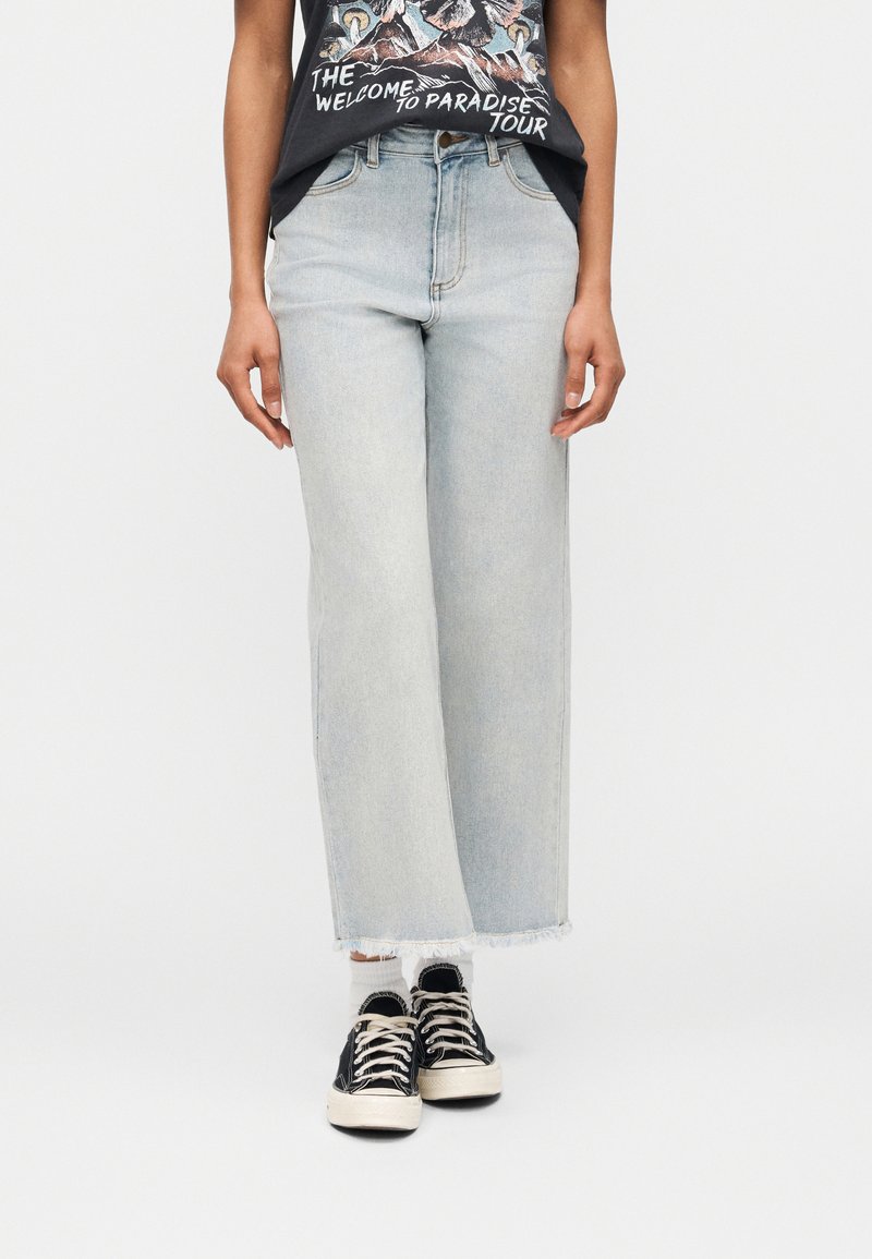 Άτομο που φοράει ανοιχτό μπλε τζιν παντελόνι με φαρδύ κόψιμο και κομμένο στο κάτω μέρος, μαύρα αθλητικά παπούτσια με λευκά κορδόνια, λευκές κάλτσες και μαύρο μπλουζάκι με σχέδιο.