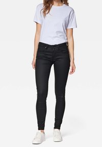 Schwarze enge Jeans mit fünf Taschen, Gürtelschlaufen und mattem Finish. Kombiniert mit einem weißen T-Shirt mit kurzen Ärmeln und weißen Turnschuhen.