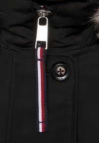 Fermeture éclair de veste noire avec une tirette argentée et un cordon en tissu rayé rouge, blanc et noir ; bouton noir avec l'inscription "HILFIGER".