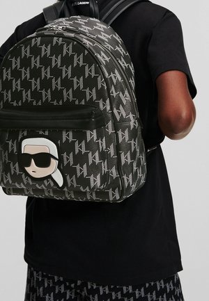 Schwarz-weiß gemusterter Rucksack mit einem Cartoon-Gesicht mit Sonnenbrille, getragen von einer Person in schwarzem Hemd und passenden gemusterten Hosen.