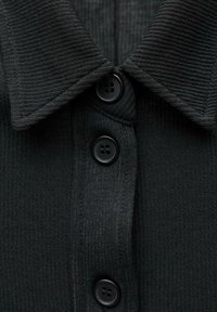 Černá žebrovaná košile na knoflíky se šířeným límcem a třemi černými knoflíky. Tkanina má texturovaný, jemný vzor.