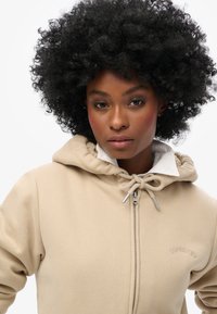 Felpa con zip beige dotata di cappuccio con cordino, tessuto testurizzato e logo ricamato. Presenta un colletto bianco interno per un contrasto.