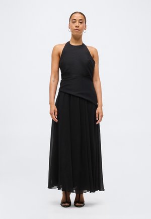 Femme debout portant un haut sans manches noir à col licou et une longue jupe noire fluide avec des escarpins noirs à bout ouvert sur un fond blanc uni.