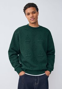Zielona bluza z podniesionymi wzorami świątecznych ozdób, z okrągłym dekoltem, ściągaczami na mankietach i dole oraz fakturowanym wykończeniem.