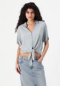 Light gray short-sleeve blouse with a tie-front design, button closure, and soft fabric paired with a light denim skirt.