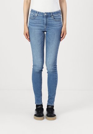 Skinny blue denim jeans with a medium wash, five pockets, and a zip fly. Worn with black sneakers featuring white stripes and gum soles.