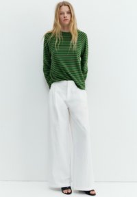 Donna con capelli biondi lunghi in piedi contro un muro bianco, indossa una maglia a maniche lunghe verde e nera a righe, pantaloni bianchi a gamba larga e sandali neri.