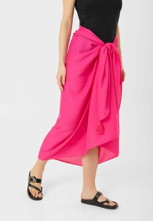 Frau trägt einen leuchtend pinken Sarong, der an der Taille gebunden ist, ein schwarzes ärmelloses Oberteil und schwarze flache Sandalen mit goldenen Schnallendetails.