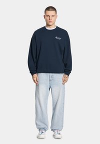 Granatowy sweatshirt z prągowymi mankietami i dołem, z małym białym logo. Noszony z jasnoniebieskimi luźnymi dżinsami i białymi trampkami.