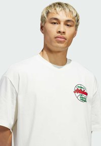 Bijela pamučna majica s obojenim grafičkim printom s tekstom i motivom košarke na lijevoj strani prsnog područja. Jednostavan dizajn s okruglim izrezom.