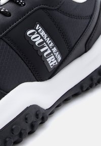 Μαύρο αθλητικό παπούτσι με υφασμένο πάνω μέρος, λευκή σόλα και εμφανές μαύ ρο λαστιχένιο λογότυπο με λευκό κείμενο στο πλάι.