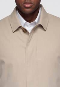 Chaqueta ligera beige con cuello con solapa, cierre con un botón redondo, textura suave y diseño limpio y minimalista.
