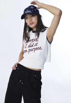 Jonge vrouw met een marineblauwe pet met patroon, wit mouwloos topje met de tekst "Oops, I did it on purpose", en zwarte broek met aantrekkoord.