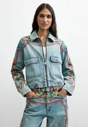 Kvinde iført lyseblå cowboyjakke og jeans med farverige blomster- og swirlmønstre, stående med hænderne i lommerne mod en ensfarvet baggrund.