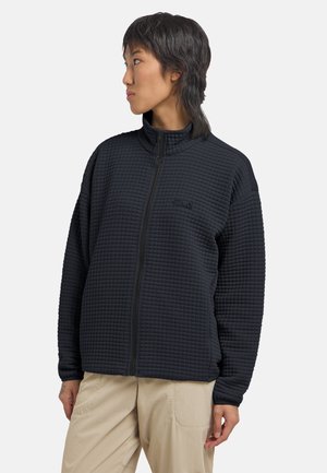 Persoon draagt een zwart doorgestikt jack met ritssluiting en een hoge kraag, en beige broek, kijkt naar links tegen een effen achtergrond.