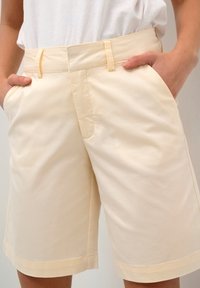 Krémové bavlněné kraťasy s přední kapsou a poutky na opasek. Mají hladkou texturu a mírně uvolněný střih, končí nad koleny.