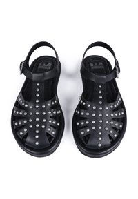 Sandalias negras con una superficie texturizada, con un diseño recortado y tachuelas metálicas, aseguradas con una correa al tobillo y hebilla.